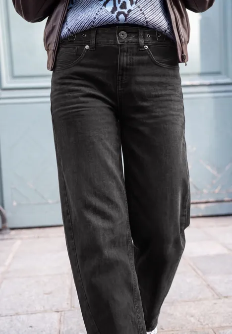 Jeans Relaxed Barrel Leg o kroju Loose Fit black washed Osoba w czarnych, prostych dżinsach i wzorzystej koszuli stoi przed niebieskimi drzwiami.