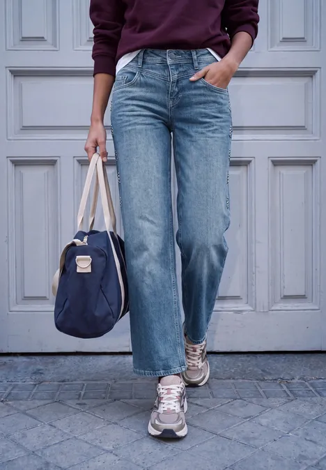 Persoon draagt wijde blauwe jeans, sportschoenen en houdt een donkerblauwe tas voor een lichte deur.