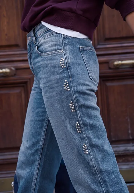 Heup in een blauwe spijkerbroek met studs langs de zijnaad, voor een houten deur.