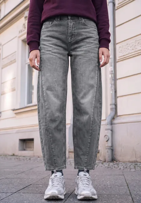 Unterkörper einer Person in grauen Jeans und weißen Sneakern auf Bürgersteig vor einer Hauswand.
