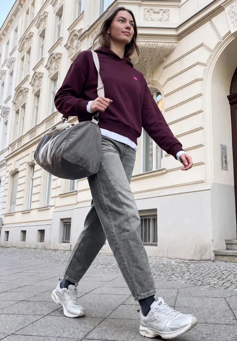 Eine junge Frau in Kapuzenpullover, Jeans und Sneakern trägt eine Sporttasche und geht eine Straße entlang.
