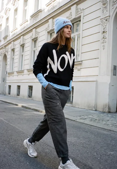 Femme en pull noir 'NOW', col roulé bleu, joggings gris et baskets blanches, passant devant une façade haussmannienne.