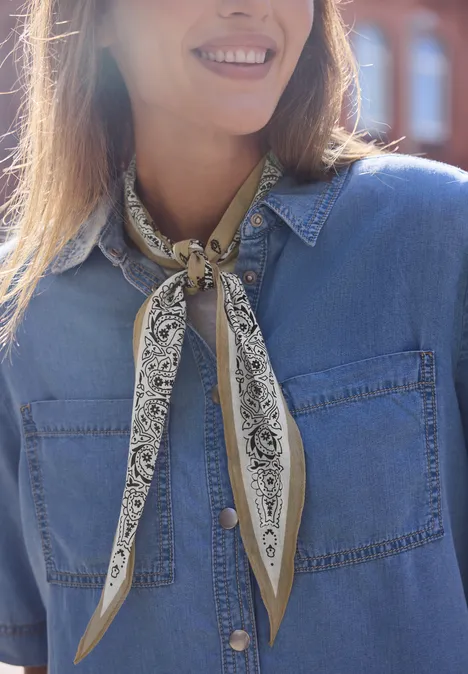 Frau mit jeansblauer Bluse und gemusterter Halstuch-Bandana, lächelnd vor einem Backsteinbau.