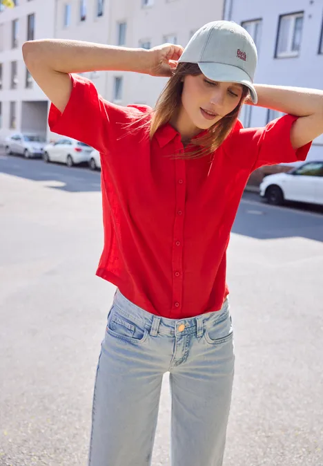 Person mit rotem Hemd und Jeans, trägt Baseballkappe, steht auf sonniger Straße und hält Hände hinter Kopf.