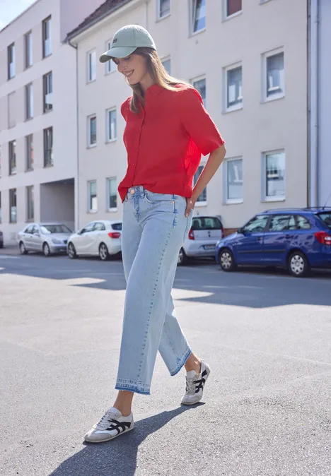 Person mit rotem Pullover, blauer Jeans und Loafers steht auf sonniger Straße mit geparkten Autos.