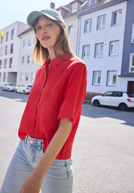 Eine Person in rotem Oberteil und Jeans steht auf einer sonnigen Straße mit Häusern im Hintergrund.