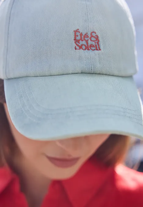 Eine Person trägt ein hellblaues Jeanscap mit roter Aufschrift 'Été & Soleil'. Gesicht unscharf.