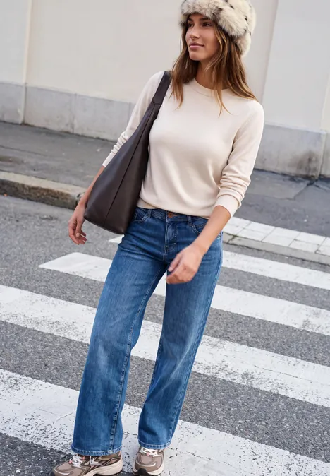 Bonnet en fausse fourrure avec motif vache smooth stone sand Eine Frau in weitem Jeans, hellrosa Pullover und Fellmütze überquert einen Zebrastreifen mit Tasche.