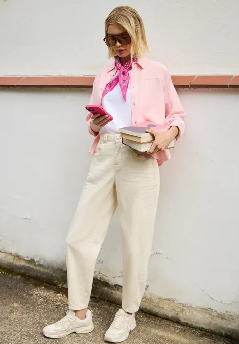 Eine junge Frau in rosa Bluse lehnt an einer Wand, hält Smartphone und Bücher in der Hand.
