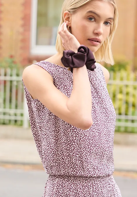 Scrunchie mit Ring-Detail very berry Frau trägt ärmellose, geblümte Bluse mit schwarzer Hals-Schleife, steht vor Zaun im Freien.
