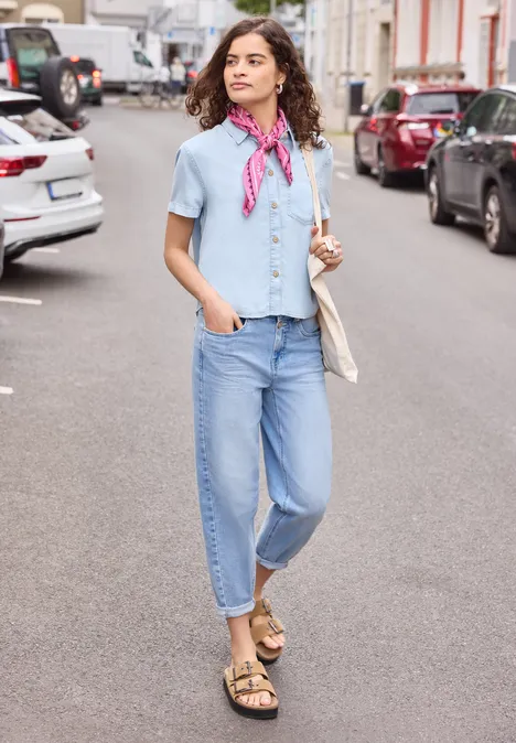Une femme marche dans une rue urbaine, portant jean, chemise bleu clair, foulard rose et sandales.