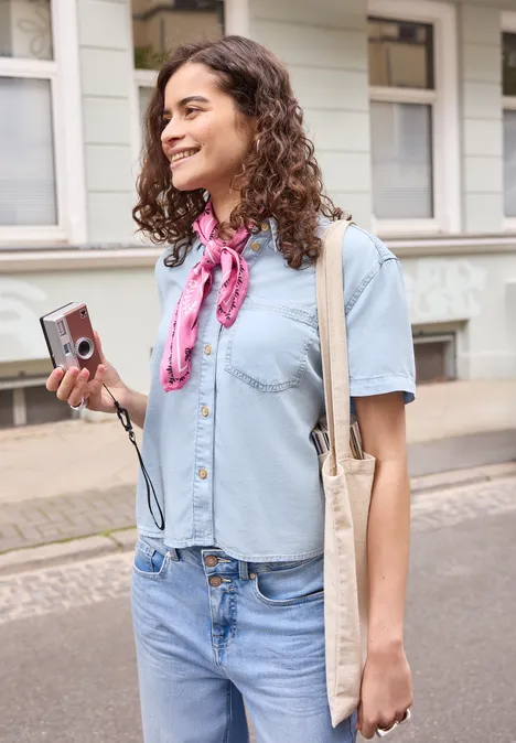Femme tenant un appareil photo, chemise bleue, jean et foulard rose en milieu urbain.