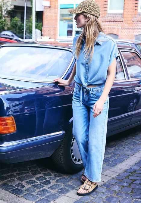 Une femme en jean, top bleu et chapeau léopard se tient près d'une voiture vintage bleue.