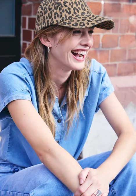 Une femme souriante en chemise en jean et casquette léopard, assise devant un mur de briques.