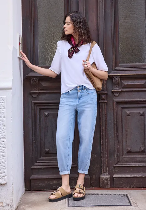 Tuch mit Mustermix multicolor Eine Frau lehnt in weißem Shirt, Jeans und Sandalen vor einer alten Holztür.
