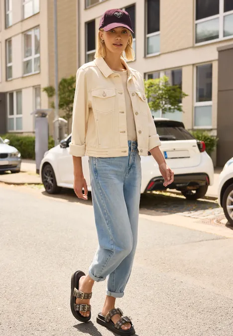 Frau in Jeans, beiger Jacke und Cap spaziert in Sandalen eine sonnige Straße entlang, lässiger Look.