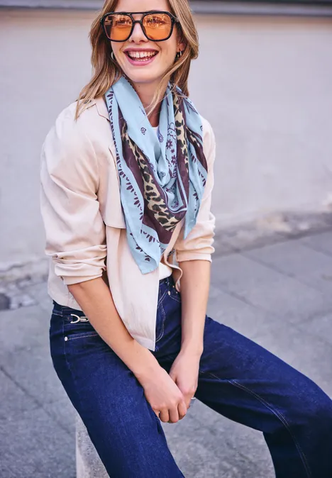 Une jeune femme souriante en lunettes de soleil, foulard et jean s’appuie contre un mur.