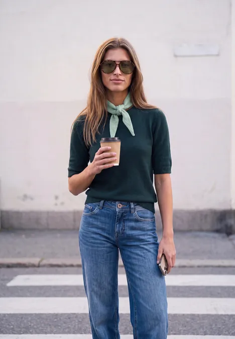 Strickschal in Diamantform grayed mint mel. Eine Frau mit Sonnenbrille, grünem Tuch um den Hals, Pullover und Jeans hält einen Kaffee und ein Handy.