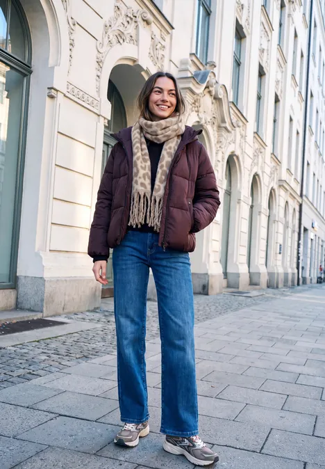 Frau in Winterjacke und Jeans geht lächelnd eine Straße mit historischen Gebäuden entlang.