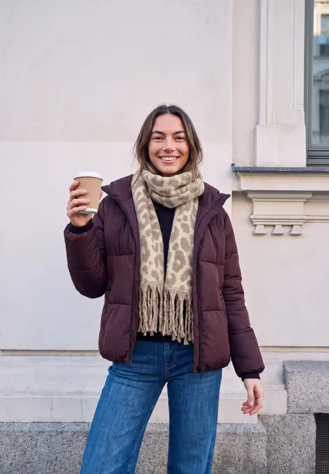 Frau steht vor Gebäude, trägt Winterjacke und Schal, hält Kaffee und lächelt in die Kamera.
