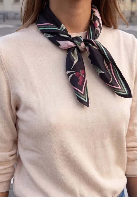 Satijnen doek met bloemmotief inked grey Vrouw draagt beige trui en zwarte zijden sjaal met roze print om haar hals geknoopt.