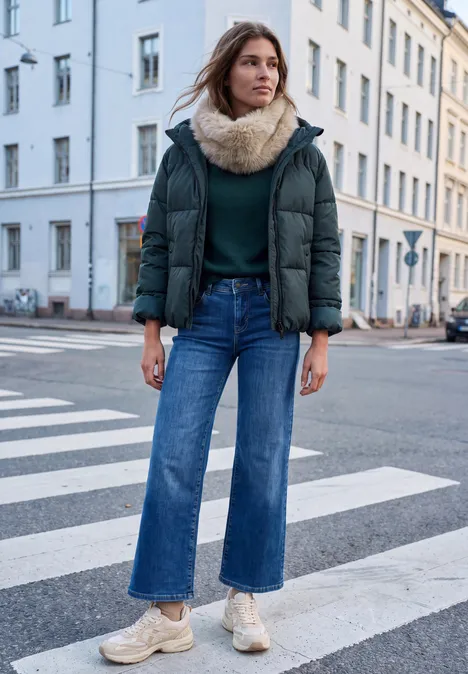 Frau im Winteroutfit mit Parka, breiter Jeans und Schal steht auf Zebrastreifen vor Häuserfassade.