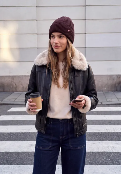 Strick-Beanie in Unifarbe russet red Junge Frau in Lederjacke mit Fellkragen und Mütze, hält Kaffee und Handy an einem Zebrastreifen.