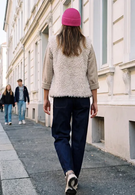 Strick-Beanie in Unifarbe ruby raspberry Frau mit pinker Mütze und Jeans läuft eine gepflasterte Straße entlang, im Hintergrund zwei Passanten.