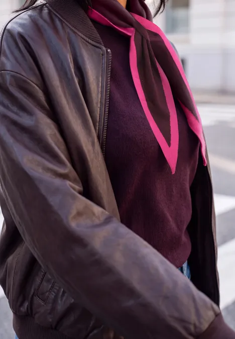 Nahaufnahme einer Person in schwarzer Lederjacke, weinrotem Pullover und pinkem Halstuch auf der Straße.