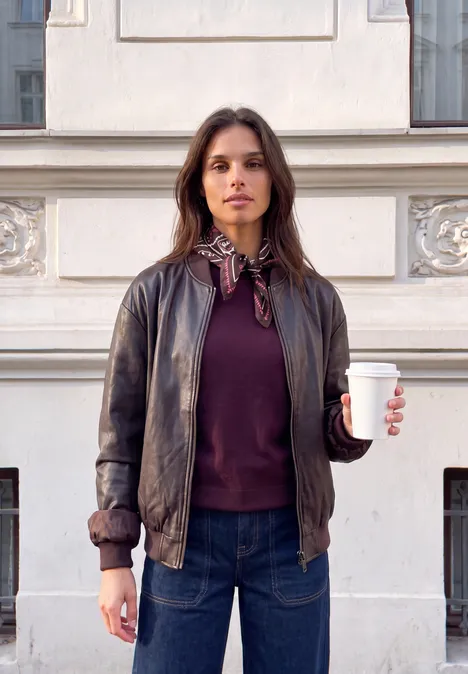 Frau trägt Lederjacke und Jeans, steht vor einem Gebäude, hält einen Kaffee zum Mitnehmen.