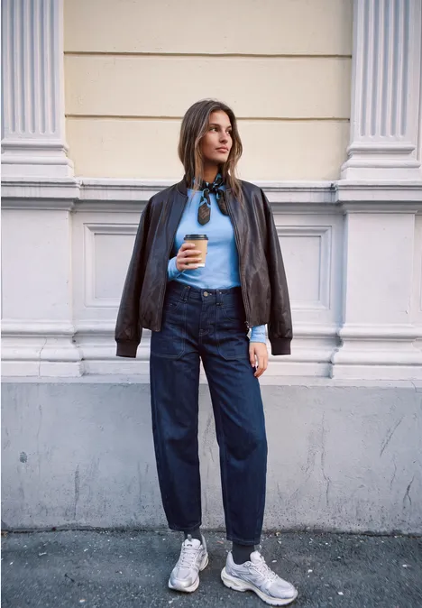 Junge Frau in Lederjacke und Jeans hält einen Kaffee vor klassischer Hausfassade.