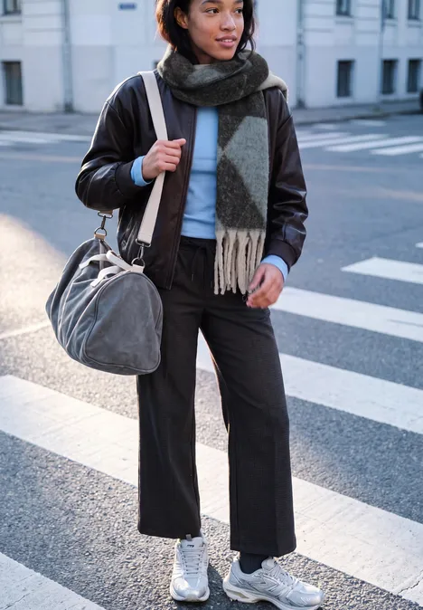 Femme en veste en cuir, écharpe et baskets blanches traversant la rue avec un sac.