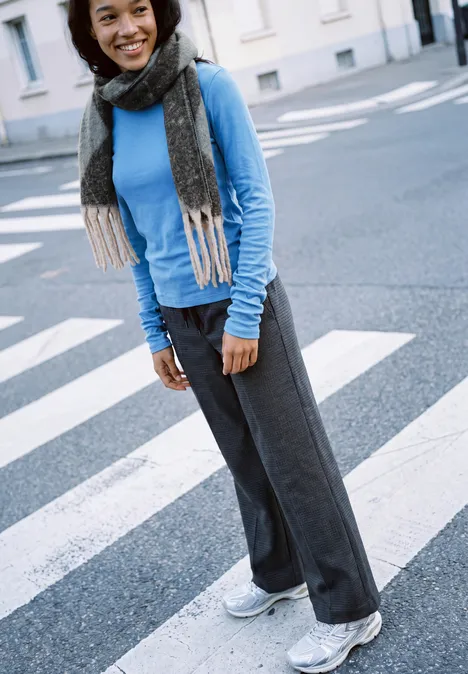 Jeune femme souriante sur un passage piéton, pull bleu, écharpe grise, pantalon ample et baskets blanches.