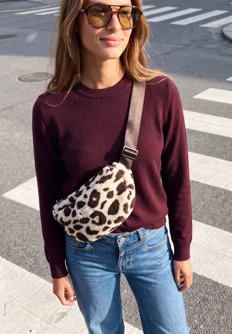 Frau mit Sonnenbrille, dunkelrotem Pullover und Jeans, trägt eine Leoparden-Bauchtasche am Zebrastreifen.