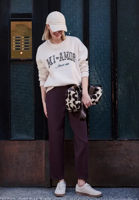 Frau mit Cap, rosa Pullover, dunkler Hose, gemusterter Tasche und weißen Sneakern vor dunkler Hauswand.