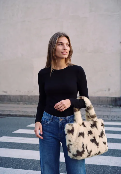 Sac cabas en fausse fourrure à motif vache smooth stone sand Junge Frau überquert Zebrastreifen in schwarzem Oberteil, Jeans und Felltasche.