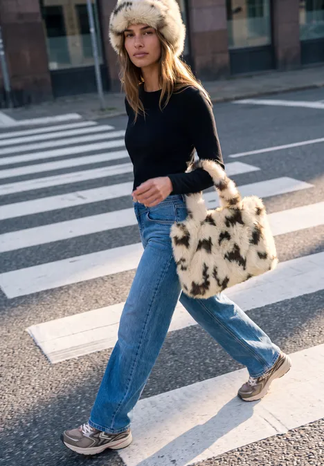 Sac cabas en fausse fourrure à motif vache smooth stone sand Frau mit Pelzmütze, schwarzem Pullover, blauen Jeans und flauschiger Tasche über Zebrastreifen.