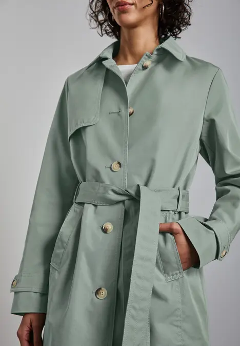 Person in grünem Trenchcoat mit Gürtel und Knöpfen, Hand in der Tasche.