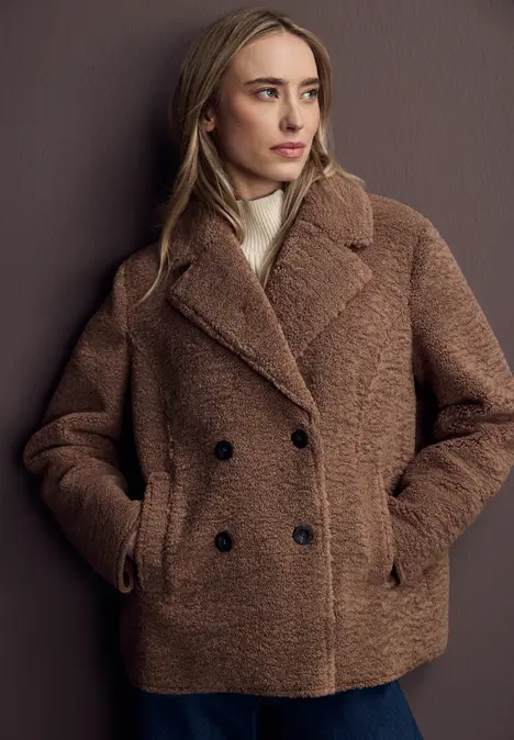 Frau mit lockigem Haar trägt braunen Teddy-Mantel, schwarzes Shirt und Jeans.