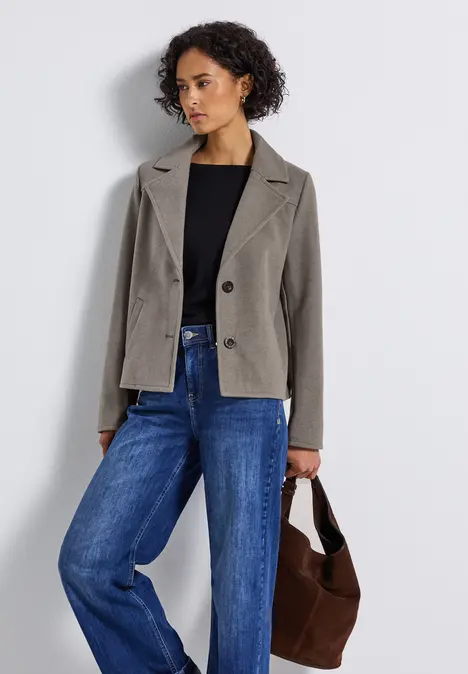 Frau mit grauer Jacke, schwarzem Shirt und blauer Jeans hält eine braune Tasche.