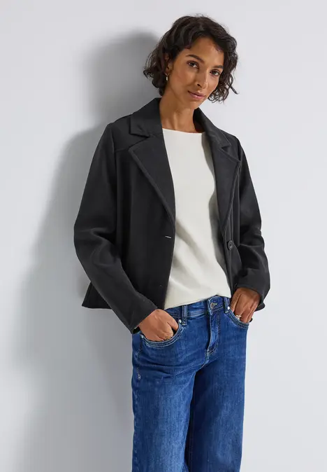 Frau mit schwarzer Jacke, weißem Shirt und blauer Jeans steht vor weißer Wand.
