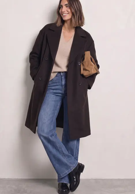 Frau in dunklem Mantel, beigem Pullover, weiten Jeans und schwarzen Stiefeln hält eine Clutch.
