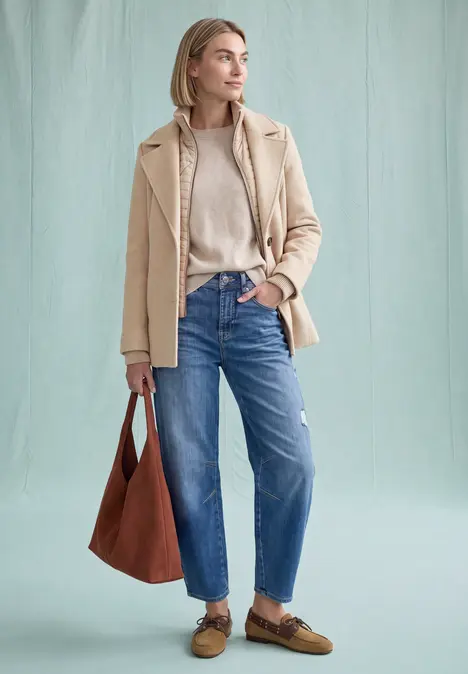 Eine Frau im hellbeigen Mantel, Pullover und blauen Jeans trägt braune Schuhe und Tasche vor hellem Hintergrund.