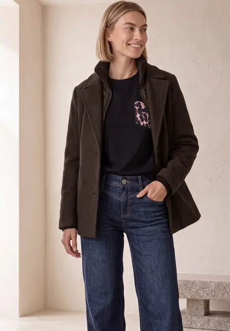 Frau in brauner Jacke, schwarzem Shirt und Jeans lächelt in hellem Raum.