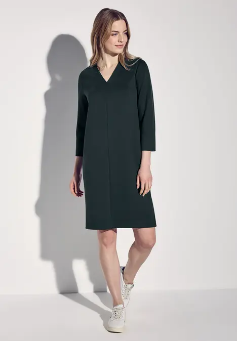 Frau in dunkelgrünem Kleid mit V-Ausschnitt und Sneakers.