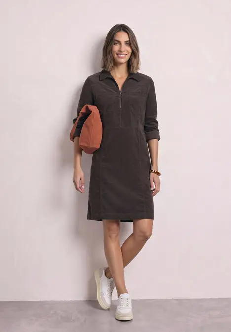Frau in dunkelbraunem Cordkleid mit halbem Reißverschluss, weißen Sneakern und orangem Clutch.