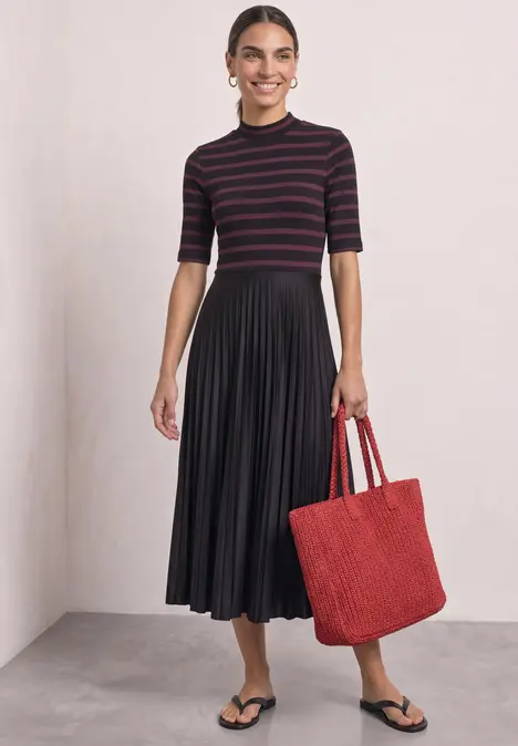 Frau in gestreiftem Oberteil, schwarzem Plisseerock, schwarzen Sandalen und rotem Shopper