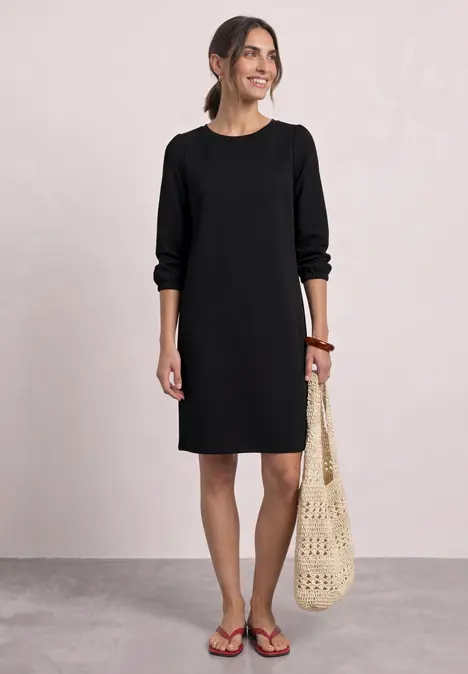 Frau im schwarzen, knielangen Kleid mit langen Ärmeln und gehäkelter Tasche, lächelnd und in Sandalen.
