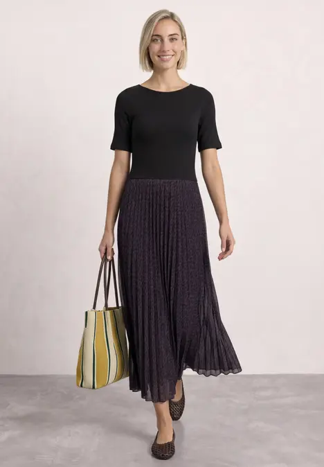 Eine Frau in schwarzem Shirt und plissiertem lila Midirock trägt schwarze Ballerinas und eine gelb-weiße Tasche.