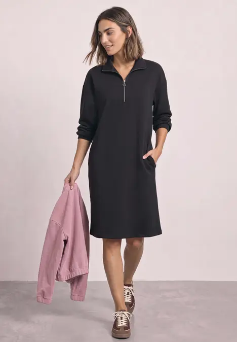 Frau im schwarzen knielangen Kapuzenkleid mit Reißverschluss hält rosa Jacke in der Hand.